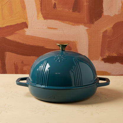 Teal enameled cast iron dome-shaped cookware on a beige surface with a colorful abstract background.