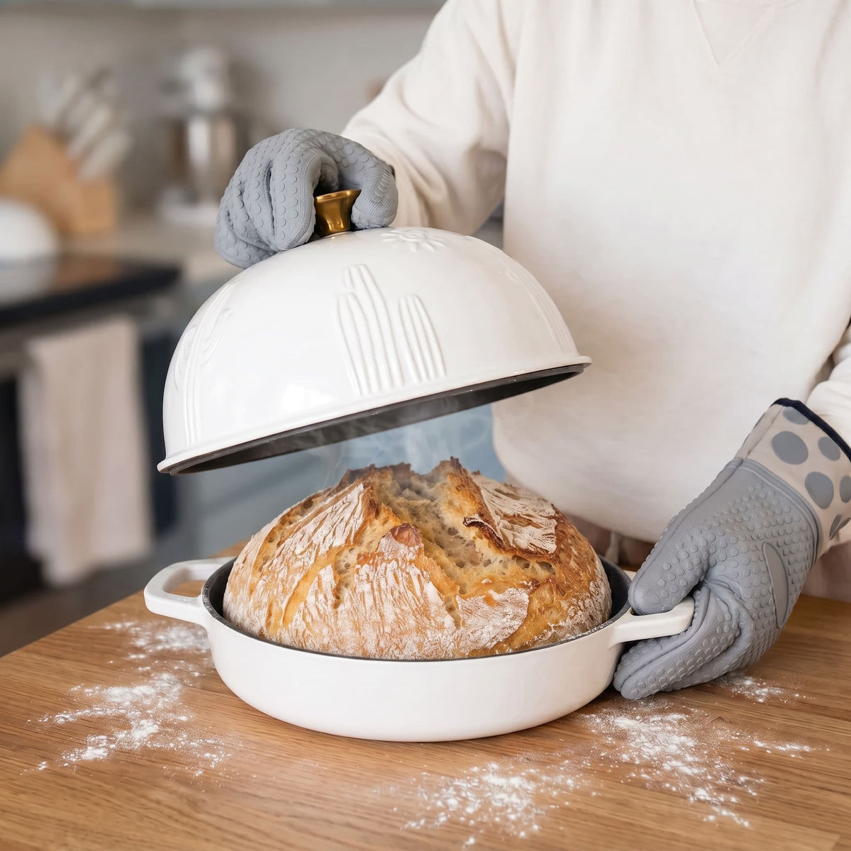 A person wearing gray oven mitts lifts the pearl white domed lid of a round white baking dish containing a rustic loaf of bread, set on a wooden table with flour scattered around.