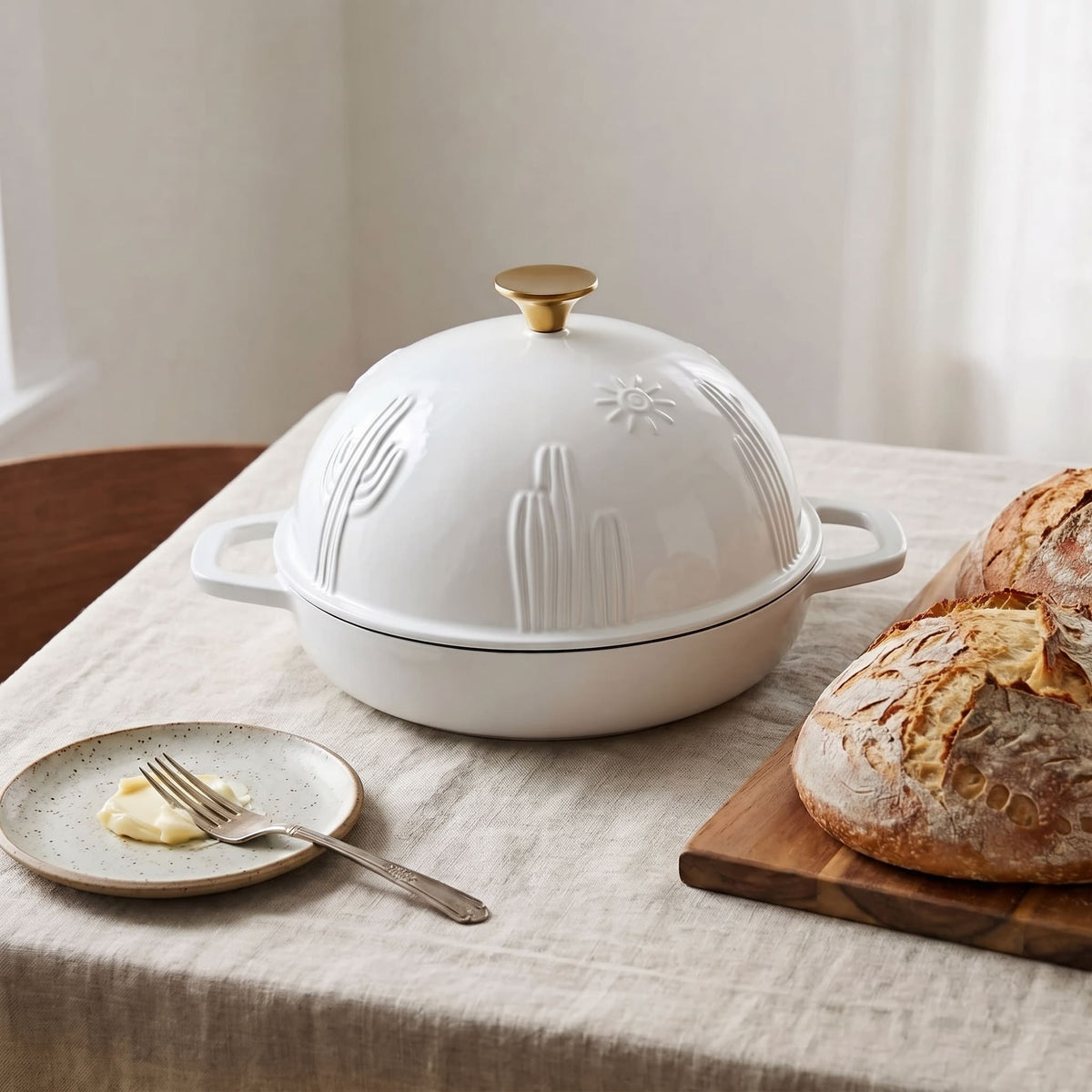 A pearl white cast iron lidded bread pan  with a gold-toned knob and embossed cactus and sun motifs, set on a linen tablecloth beside a plate with butter and fork and a loaf of bread on a wooden board.