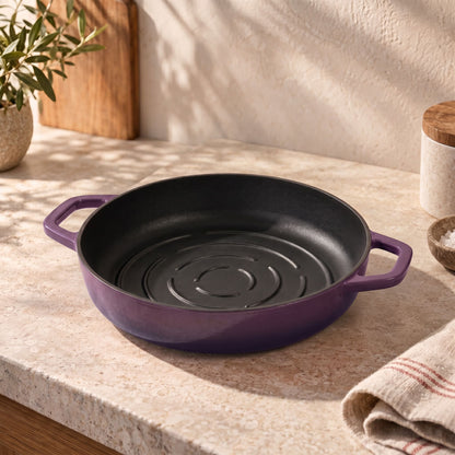 A round cast iron bread pan with a plum purple exterior and black interior, resting on a beige stone countertop in a warm kitchen setting.