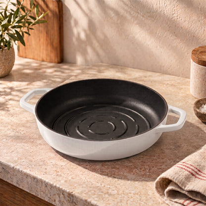 A pearl white enameled cast iron bread pan with a black interior sits on a beige stone countertop in a warm, sunlit kitchen setting.