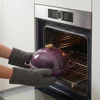 A plum purple enameled cast iron Dutch oven with a lid is being pulled out of a built-in oven using gray quilted oven mitts. The oven is stainless steel with a dark glass door.