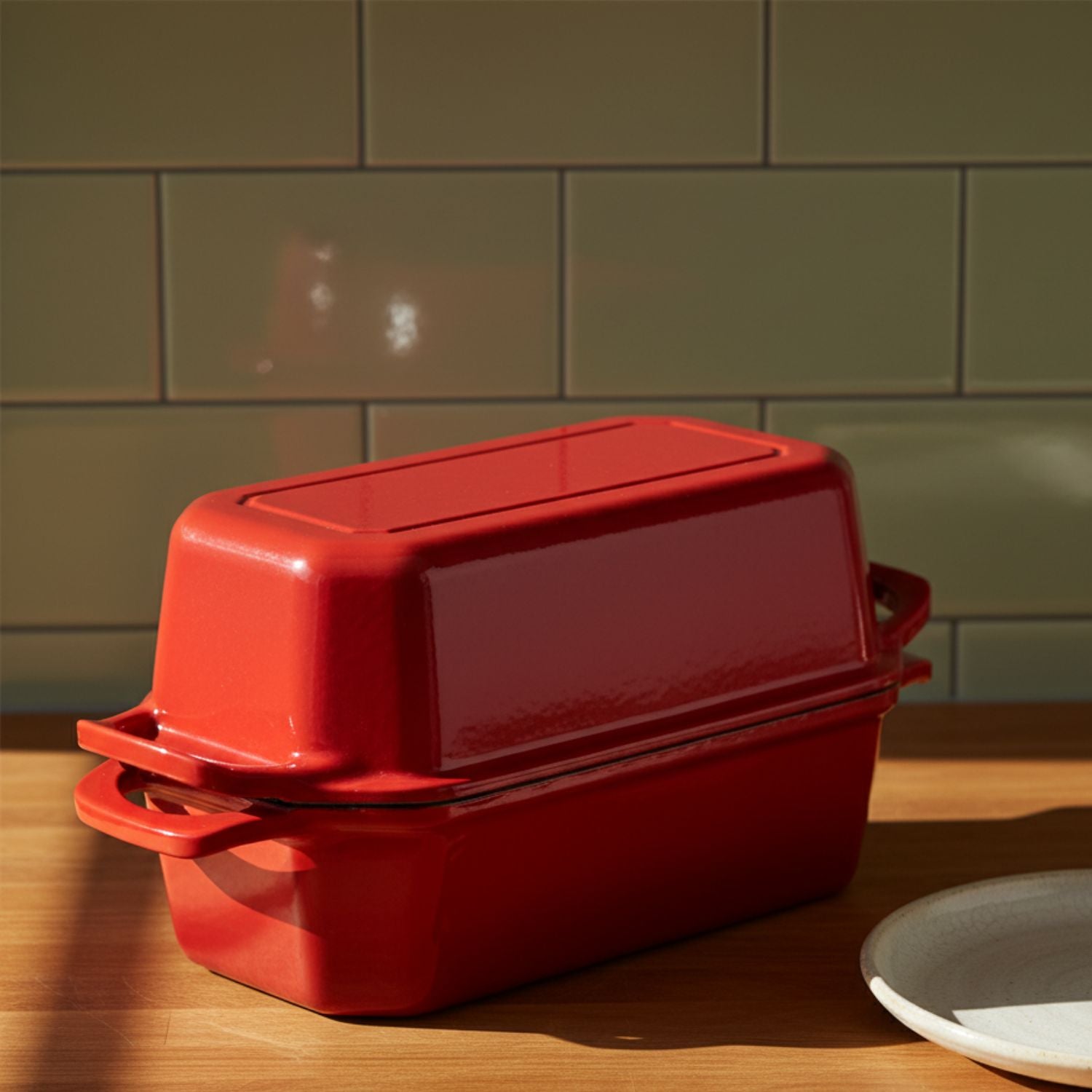 Red cast iron bread pan nz baking dish with lid on a wooden surface against a green tiled wall.